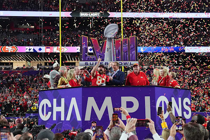 Travis Kelce points up while standing with Andy Reid and other Chiefs members in front of a Super Bowl LVIII sign and on top of a “Champions” platform